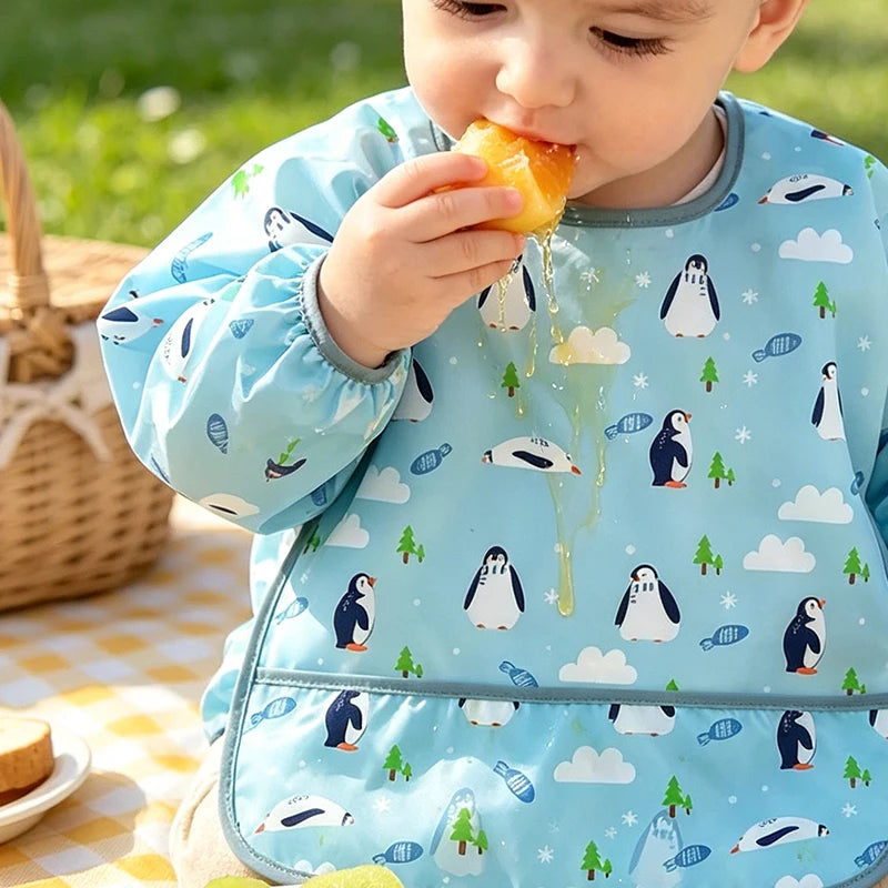 A baby is eating food while wearing a light blue waterproof bib featuring a cartoon design of penguins and clouds.