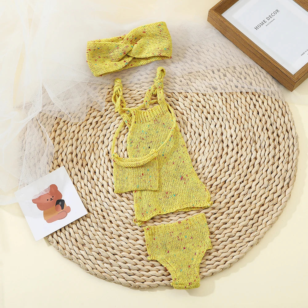 A yellow knitted doll outfit set for a baby, including a top, shorts, and a bow, displayed on a woven mat.