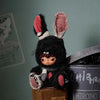 A black plush doll with pink ears and a hood sits on a wooden box next to a gray container.