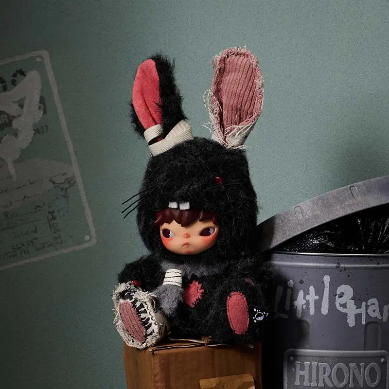 A black plush doll with pink ears and a hood sits on a wooden box next to a gray container.