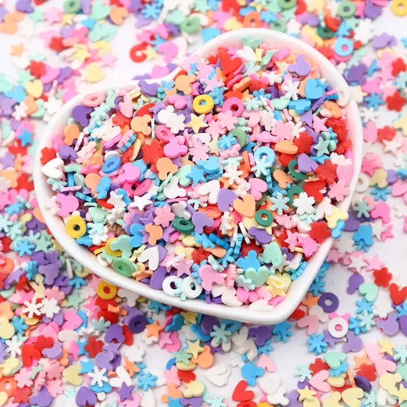 A heart-shaped bowl filled with colorful polymer clay sprinkles for slime and craft decor scattered on a white surface.