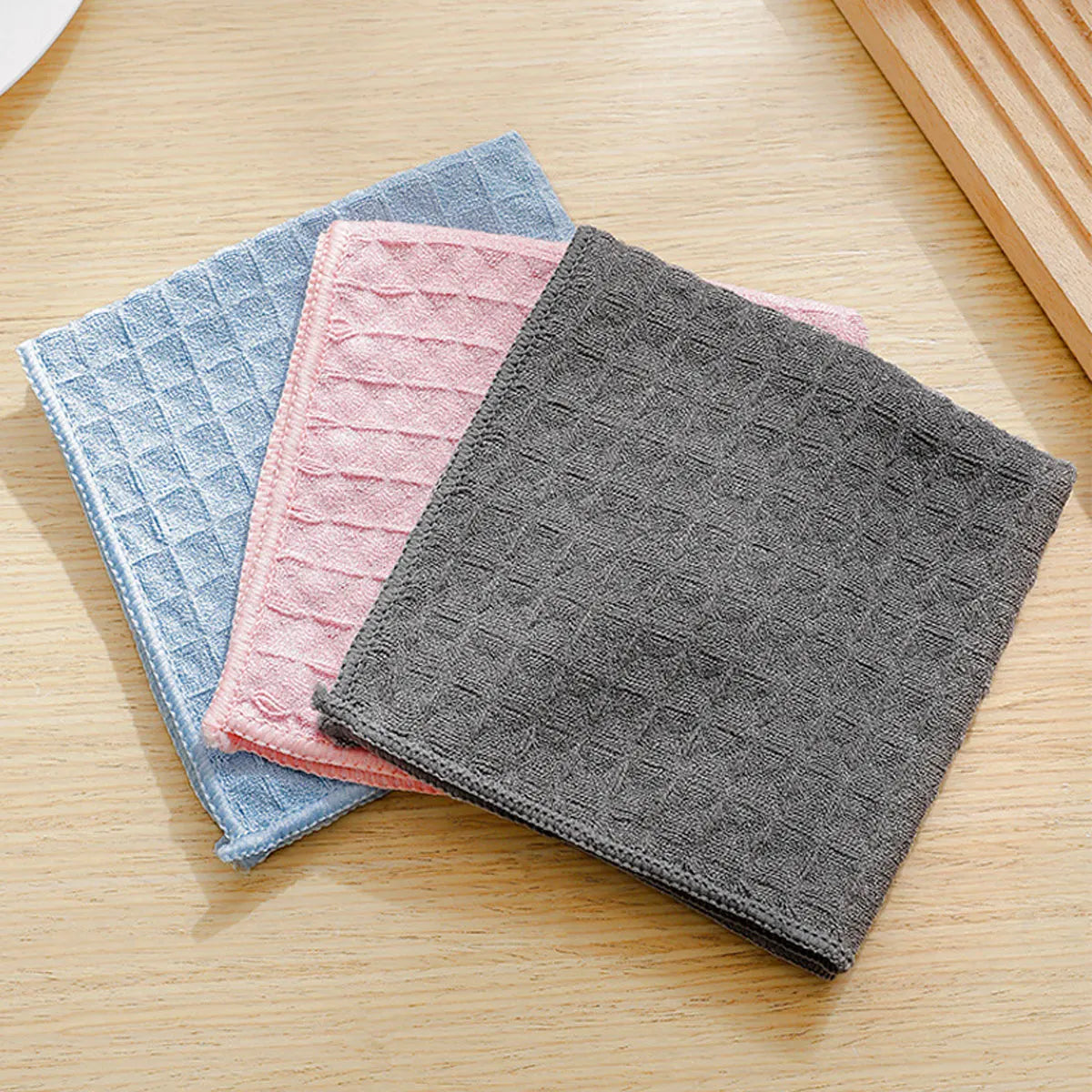 Three stacked microfiber kitchen cleaning cloths in blue, pink, and gray on a wooden surface.