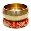 A brass singing bowl resting on a red fabric cushion with gold embroidery.