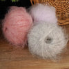 Three balls of long-haired alpaca blend yarn in shades of pink, lavender, and gray resting on a wooden surface.