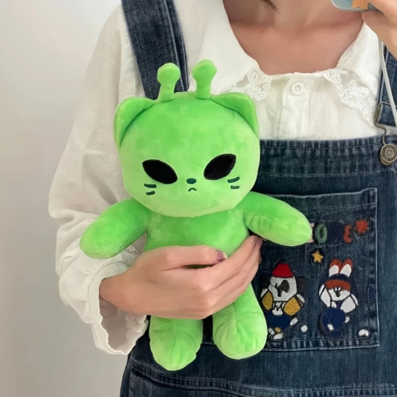 A bright green plush toy resembling an alien cat is held by a person wearing a denim apron over a white shirt.
