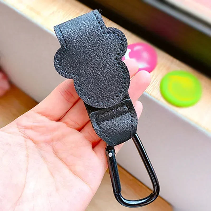 A hand holds a black PU leather stroller organizer hook with a metal loop.