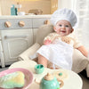 A baby wearing a chef hat and apron sits in a high chair with play food items.