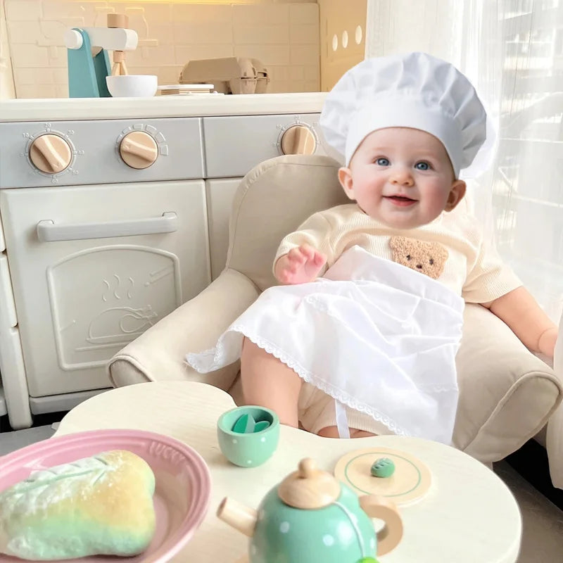 A baby wearing a chef hat and apron sits in a high chair with play food items.