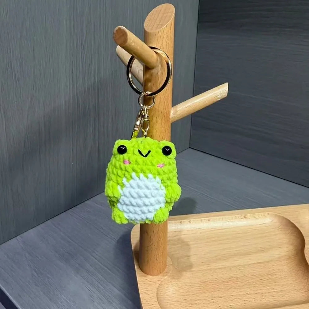 A bright green crochet frog charm with a white belly is hanging from a wooden rod and attached to a key ring.