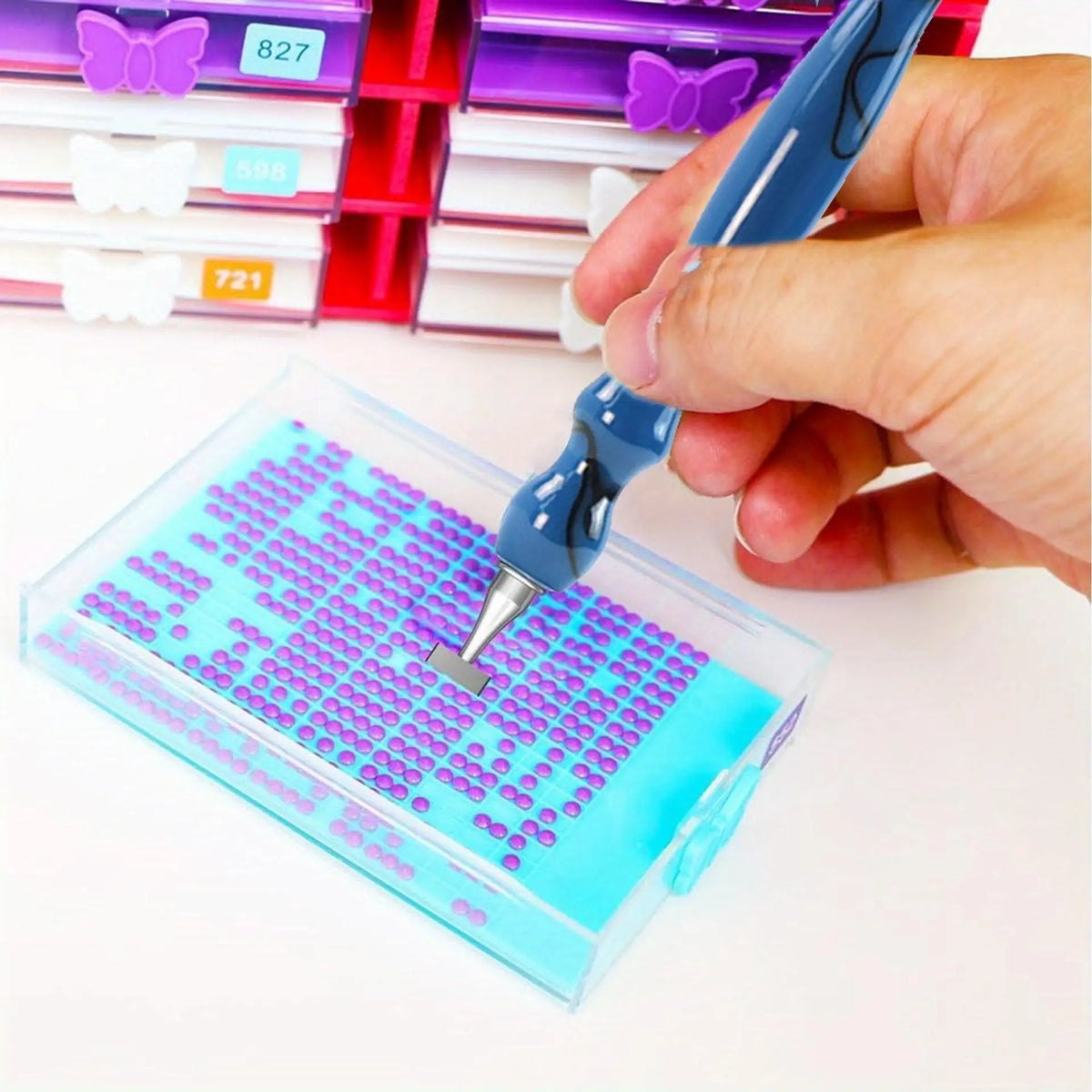 A hand holds a blue diamond art pen with an interchangeable tip, poised over a clear plastic box containing a grid of small p