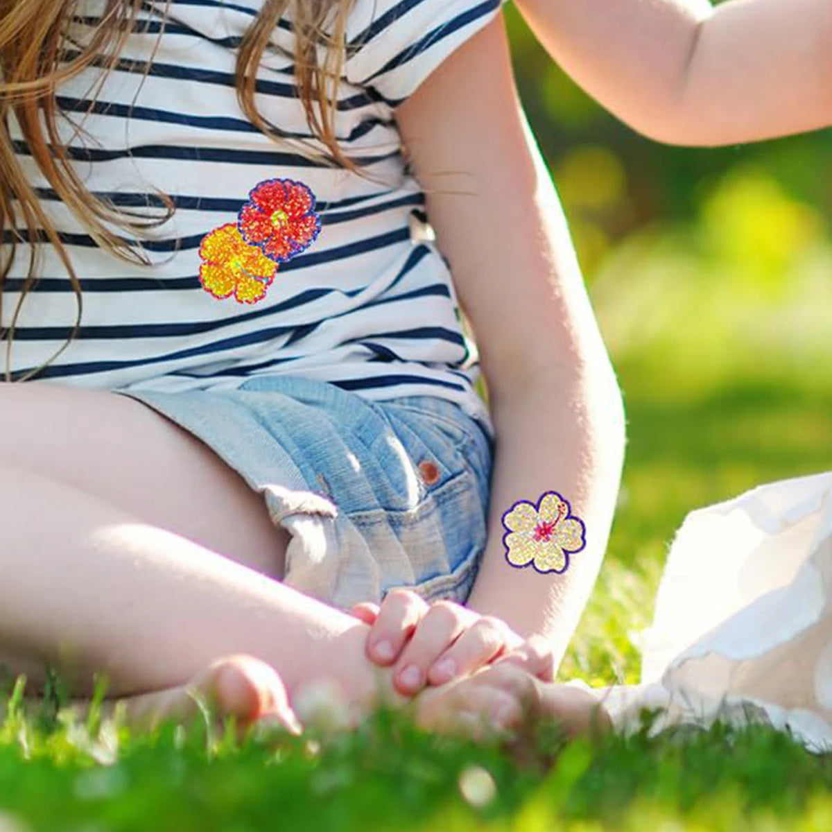 A young girl is sitting in the grass with her hands clasped, wearing a striped shirt and jeans, while holding glitter flower 