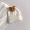 A cream knitted baby hat with a brown bear ear decoration resting on a white ribbed fabric.