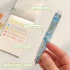 A hand holds a clear gel pen with a light blue body and a transparent barrel, next to a piece of paper showing writing exampl