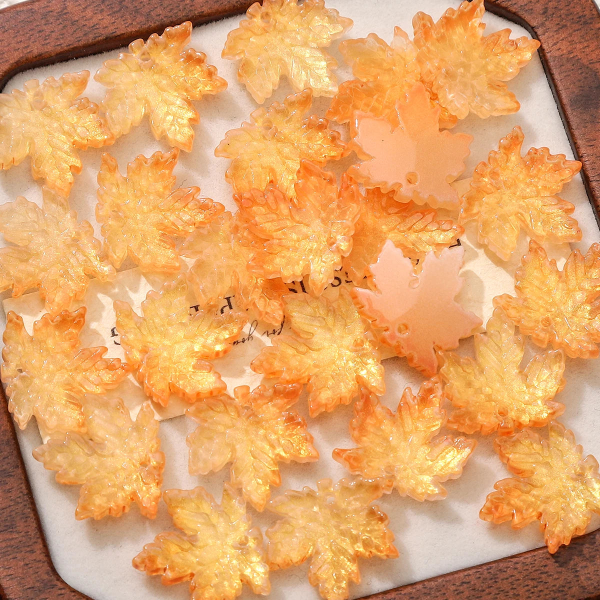 A collection of translucent orange maple leaf glass beads arranged on a wooden tray.