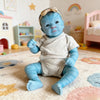 A handcrafted reborn baby doll with blue skin and features, wearing a light gray outfit, sitting on a fluffy rug.
