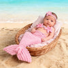 A newborn baby wearing a pink knit mermaid tail skirt and a matching headband is resting in a wicker basket on a sandy beach.