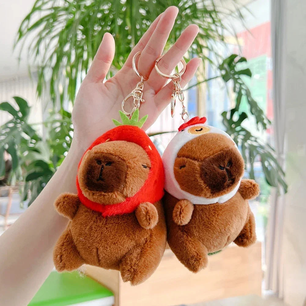A hand holds an adorable capybara plush keychain charm with a red hat and green leaves.