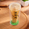 A clear container holds a set of traditional bamboo matcha whisks with Japanese text on the handle, resting on a wooden surfa