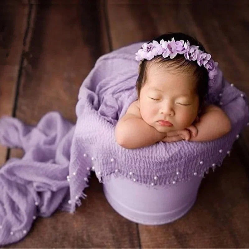 A newborn baby is sleeping wrapped in a purple knit wrap with a pearl headband.