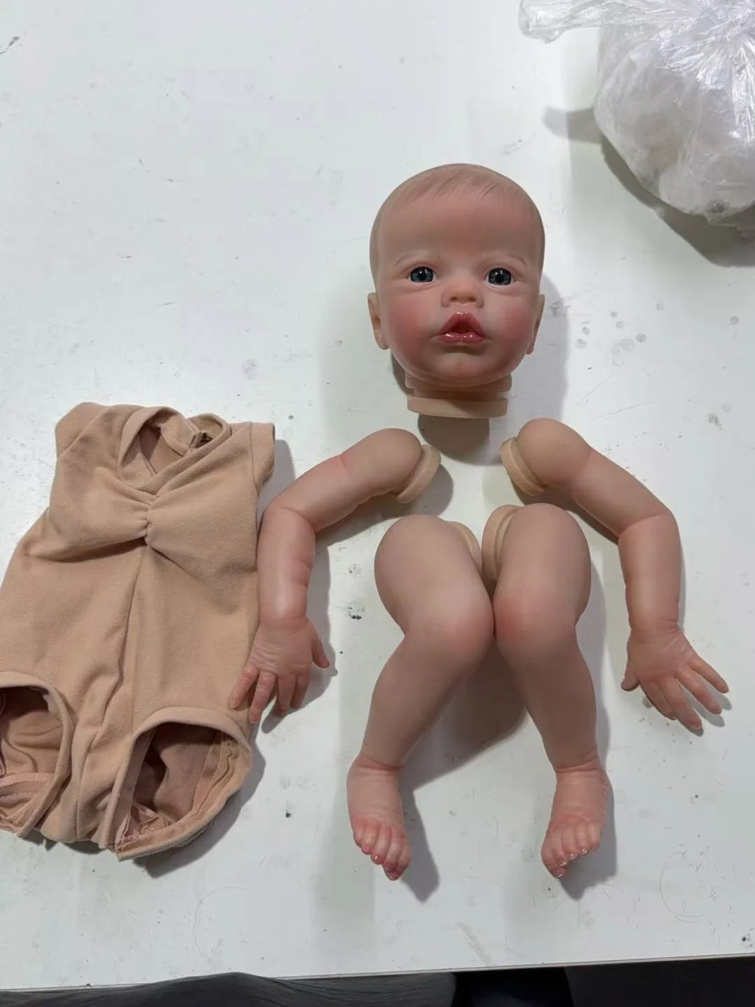 A realistic reborn baby doll kit with painted details and a cloth body is displayed on a white surface.