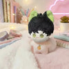 A cute anime plush keychain charm with black hair, green ears, and a light pink body resting on a fluffy surface.