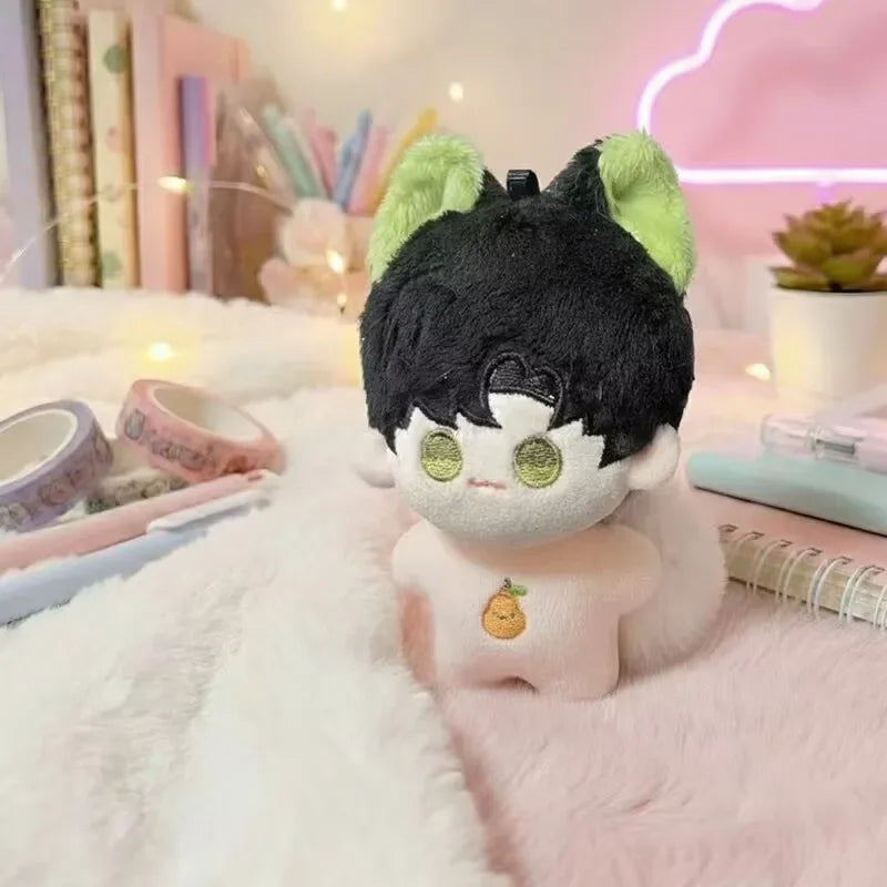 A cute anime plush keychain charm with black hair, green ears, and a light pink body resting on a fluffy surface.