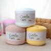 Three spools of 100g Cotton Yarn in white, pink, and yellow stacked on a wooden surface.