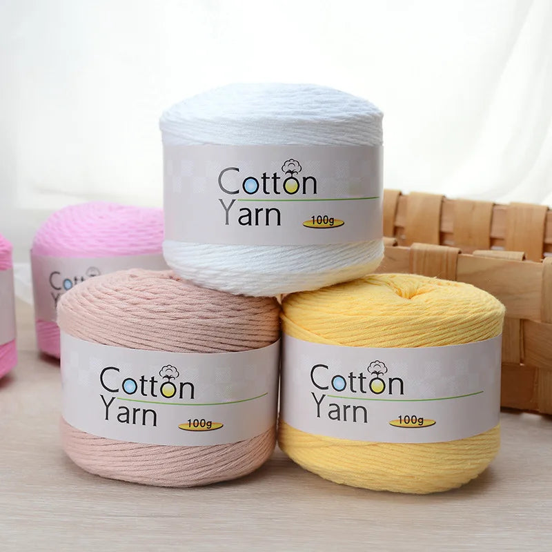 Three spools of 100g Cotton Yarn in white, pink, and yellow stacked on a wooden surface.