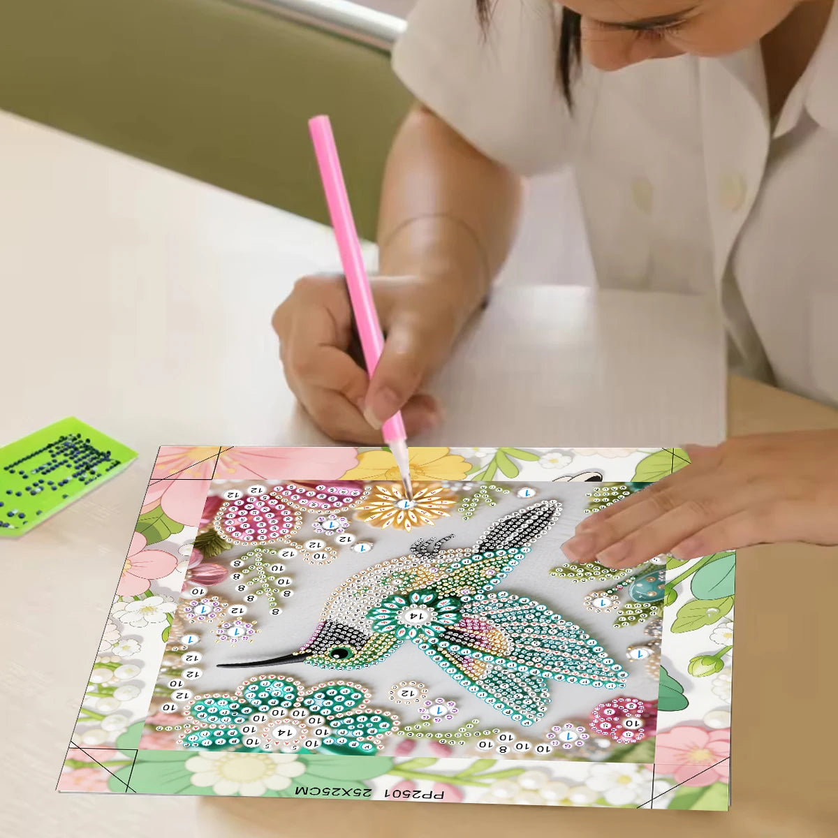 A person is painting a bird design on a diamond painting canvas using a pink tool.