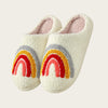 A pair of white plush indoor slippers decorated with a rainbow design in red, yellow, and gray.