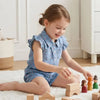 A young girl is sitting on a white rug playing with wooden building blocks and wearing a floral print sleeveless shirt and sh