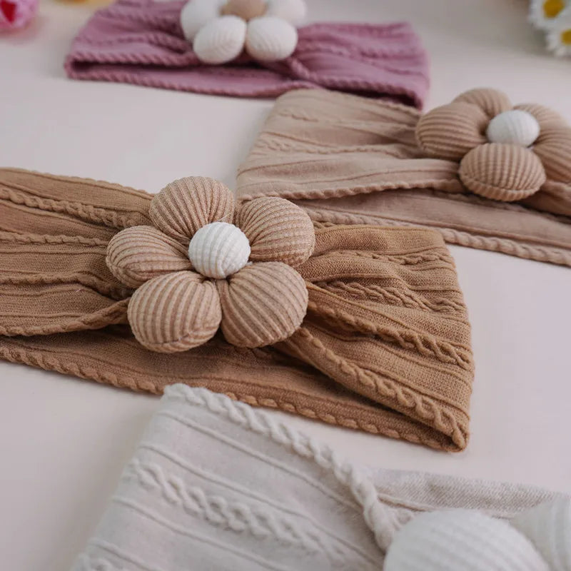 A close-up of knitted floral elastic baby headbands in beige and white on a light pink background.