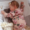 A newborn doll with soft silicone body wearing a floral pink outfit is sleeping on white fabric.