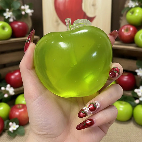 A hand holds a bright green, translucent apple-shaped sensory fidget ball with a stem against a blurred background of red and