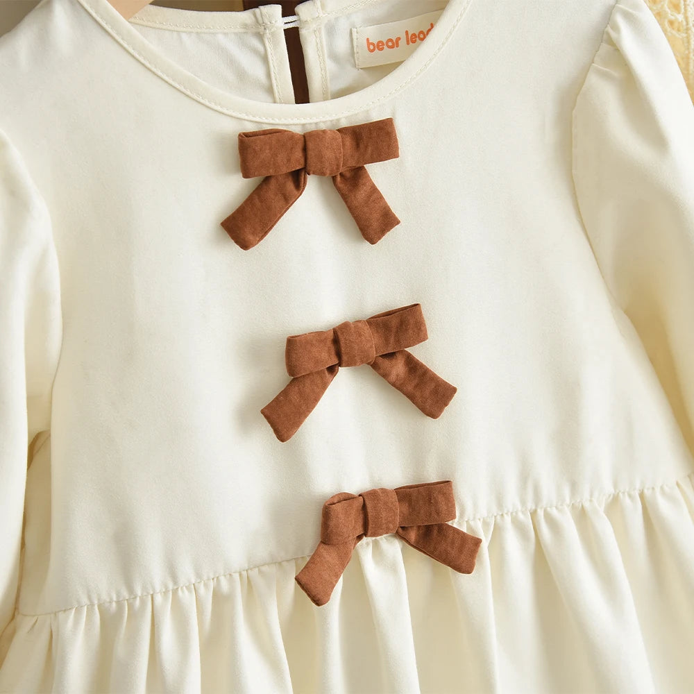 A cream-colored girls' long sleeve dress with three brown fabric bows on the chest.