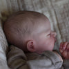 A close-up of a realistic silicone reborn doll with soft skin and light brown hair resting in a textured fabric.