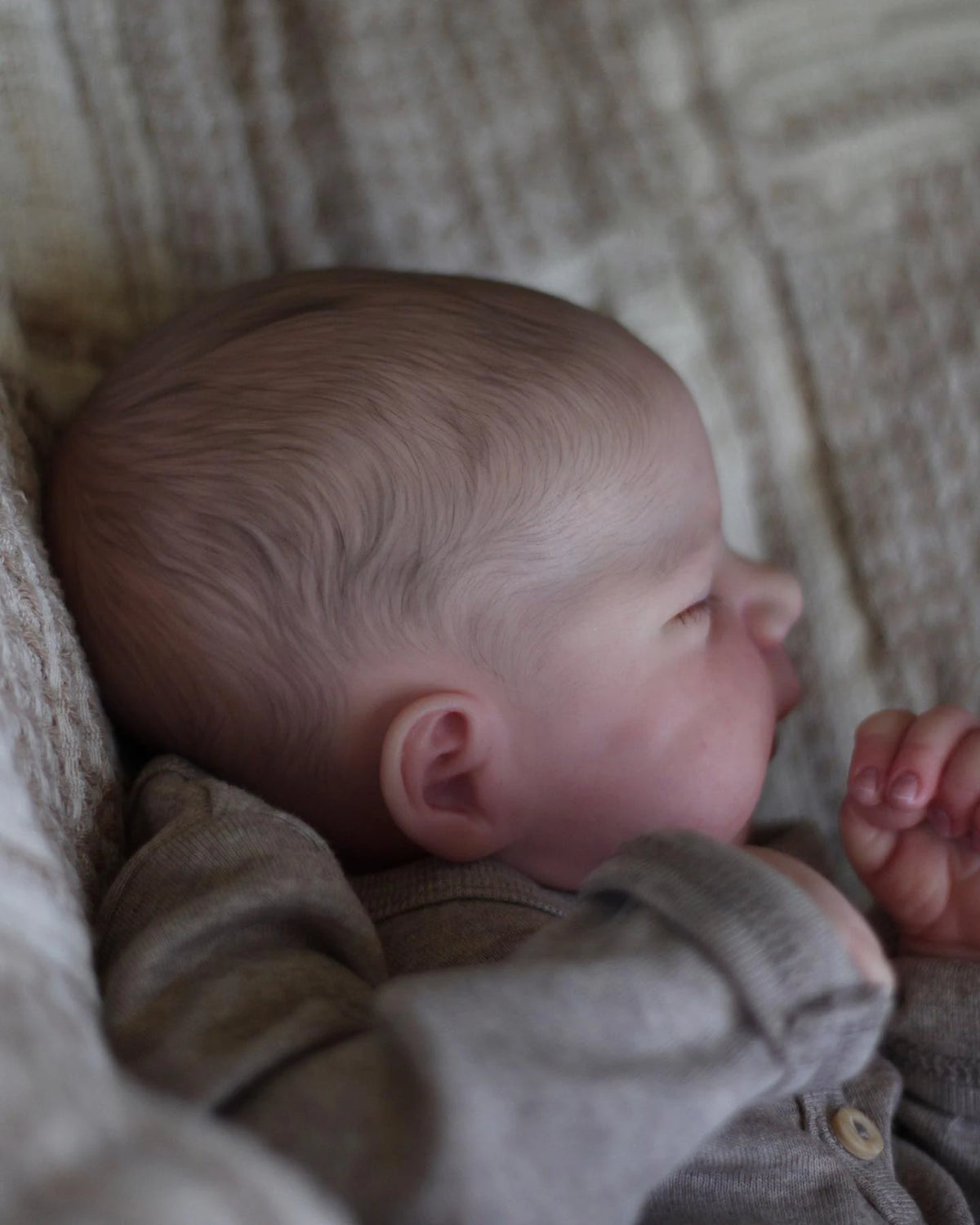 A close-up of a realistic silicone reborn doll with soft skin and light brown hair resting in a textured fabric.