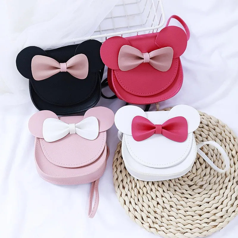 Pink, black, and white kids' hard shell crossbody bags with mouse ears and bows displayed on a woven mat.