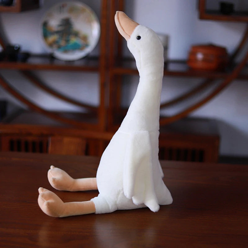 A white long neck goose plush doll with orange feet is resting on a wooden surface.