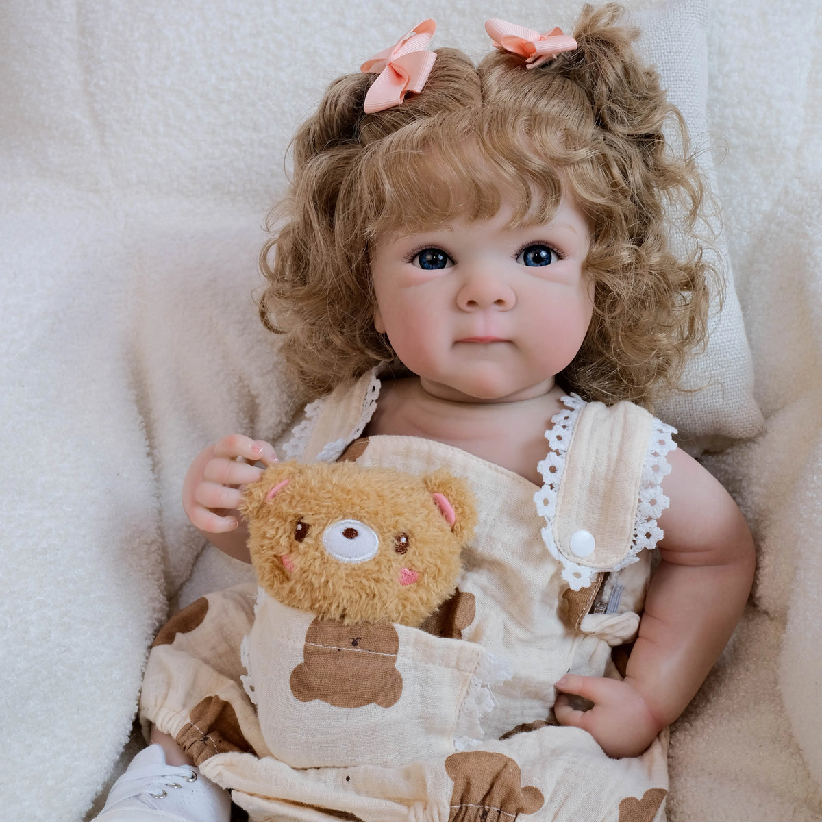 A baby doll with hand-rooted blonde hair wearing a beige outfit and sitting in a soft, light-colored fabric.