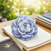 A light blue reusable flower silicone mold resting on an open notebook with a blurred green background.