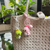 A handmade crochet dinosaur keychain with pink and yellow accents hanging from a beige woven bag.