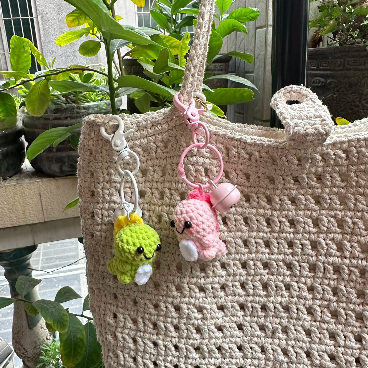A handmade crochet dinosaur keychain with pink and yellow accents hanging from a beige woven bag.