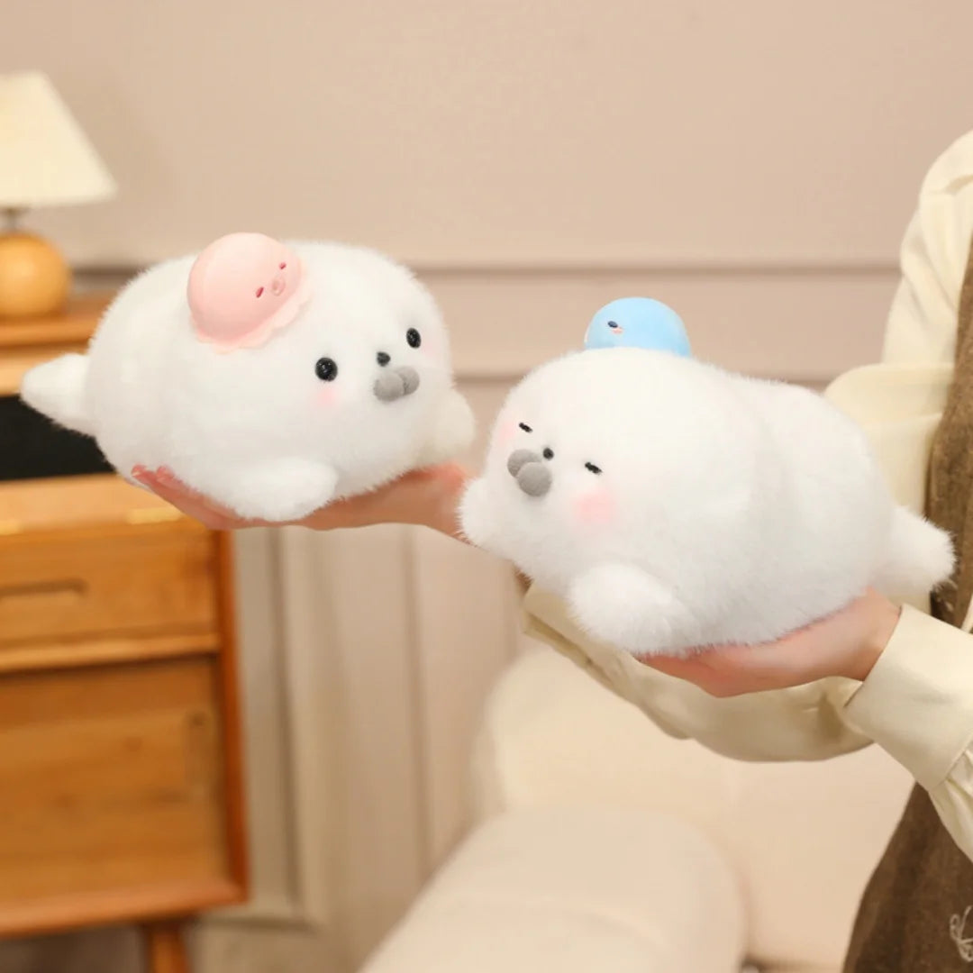 A person is holding two white seal plush toys, one with a pink hat and the other with a blue hat.