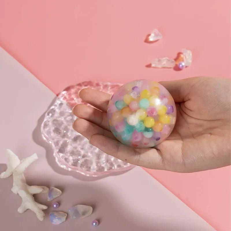 A hand holds a clear pink squishy toy filled with colorful glitter and small translucent stones on a pink surface.