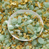 A pile of light green imitation jade charms and beads mixed with gold-colored jewelry findings on a white dish.