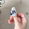 A hand holds a white figurine shaped like a blindfolded cat, featuring a small gray cat figure inside.