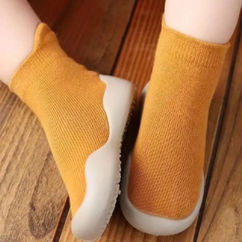 A pair of mustard yellow baby first walker socks with white soles resting on a wooden surface.