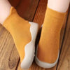 A pair of mustard yellow baby first walker socks with white soles resting on a wooden surface.