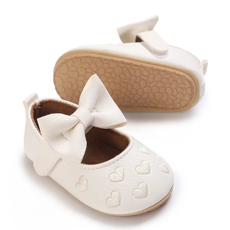 A pair of white lace-trimmed baby girl moccasin shoes with a bow and a tan sole.